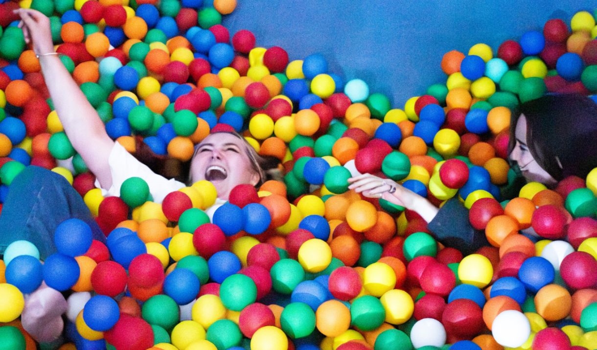 Two adults laugh and play in a giant ball pit filled with brightly coloured balls inside an indoor play area. One person raises an arm in excitement w