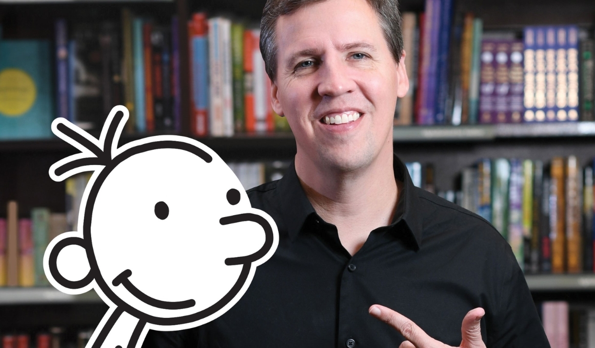 Author Jeff Kinney stands in front of a bookshelf, with a cut-out of the character Greg Heffley in front of him.