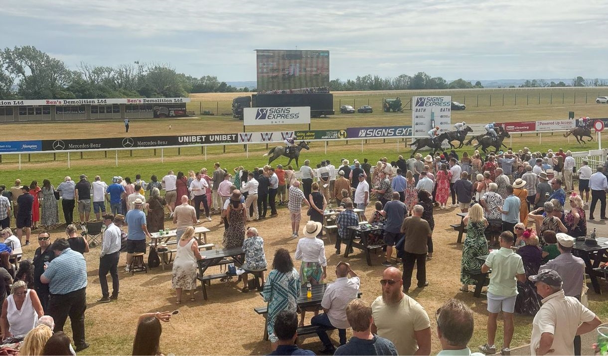 A group of people cheering and enjoying the 2025 Charity Race Day at Bath Racecourse, with a blue sunny sky.