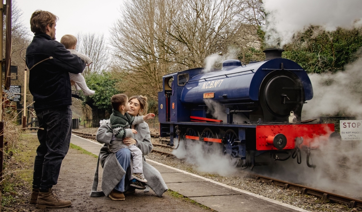 A family of four can be seen from behind waving at a blue steam train from the station platform.