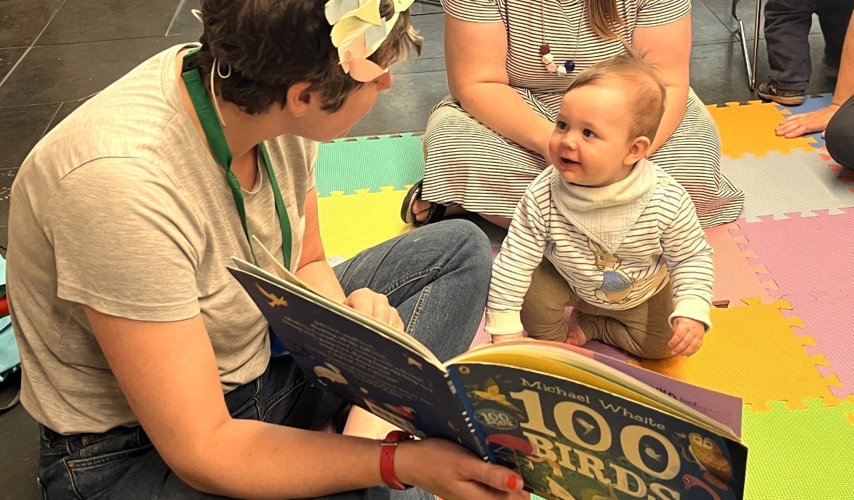 A woman reads a picture book to a baby, who is sat on a soft coloured mat
