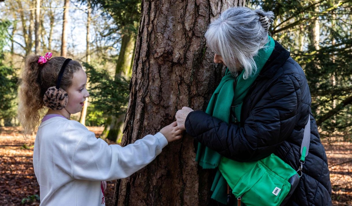 young girl and woman lean against tree trunk touching the tree bark