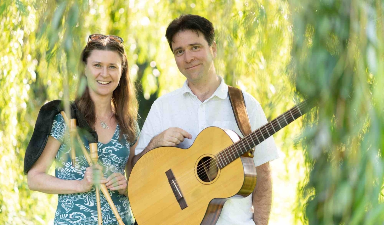 Two people sat holding musical instruments under a tree.