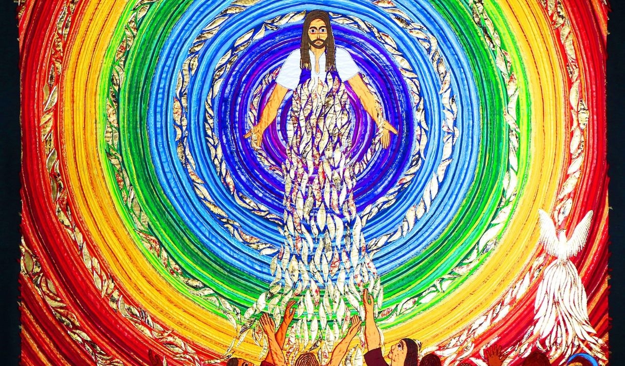 A brightly coloured embroidered panel of Jesus, surrounded by concentric circles of rainbow colours, with a crowd of people in front of Him