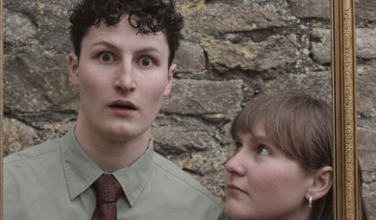 Against a brick wall, 2 individuals peer through a gold picture frame that they are holding. Sparkfest logo in corner