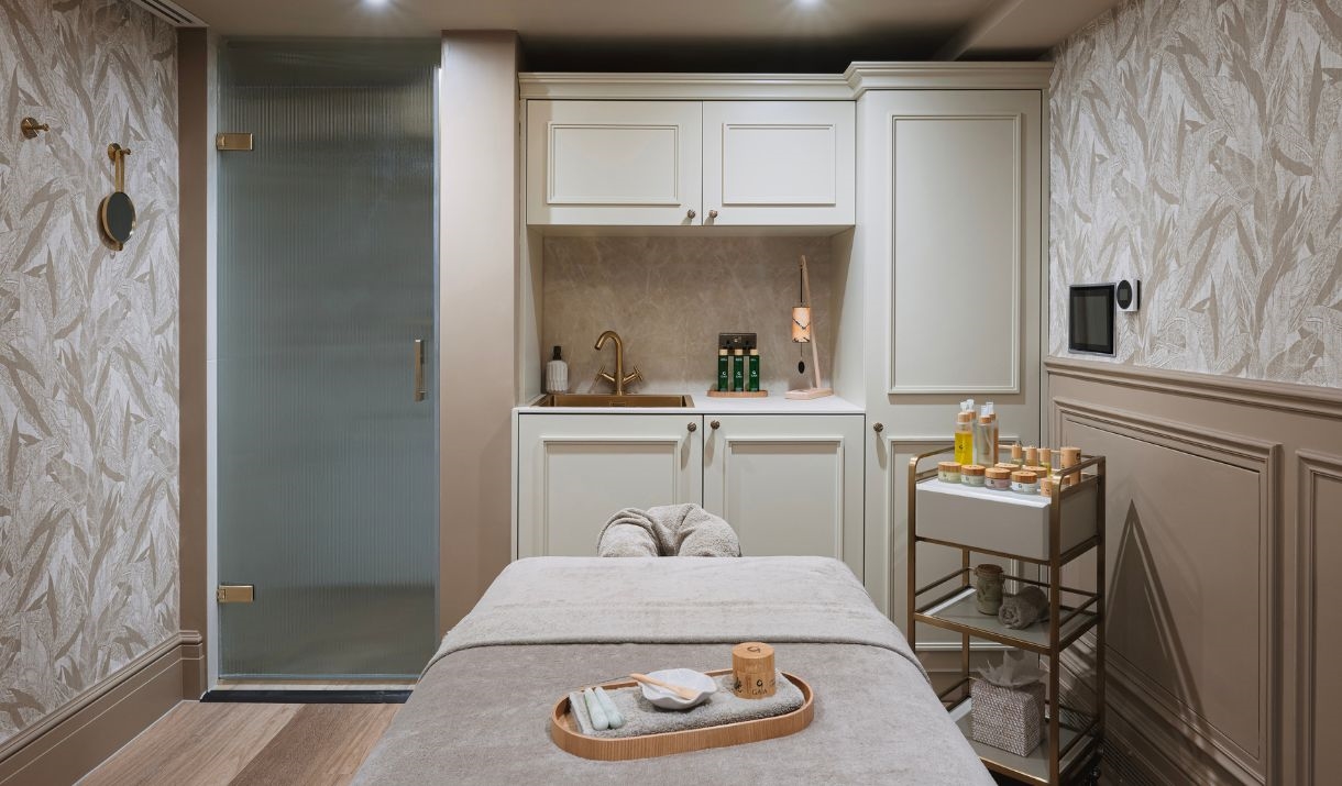 A treatment room at Francis Hotel Spa with a bed in the centre and a trolley of products to the side. The room has beige and white wallpaper with a mo