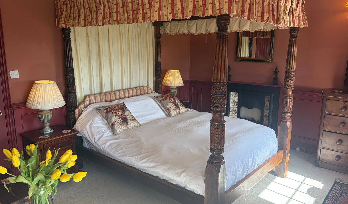 A bedroom with a wooden four poster bed with short frill curtains around the top and full length white fabric at the head of the bed.
