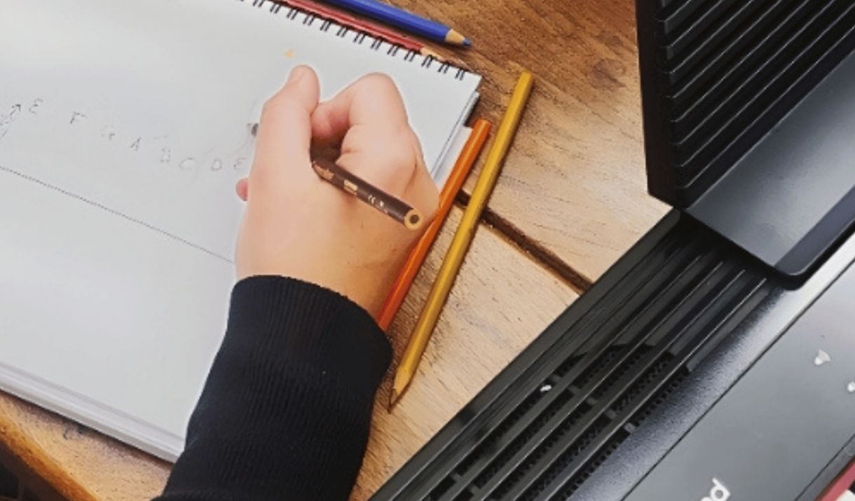 On a table there is a spread of pencils, someone writing on a notebook with a keyboard next to them,  as well as a red mug that reads "Keep calm and c