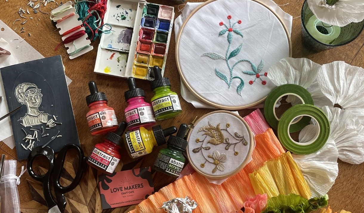 A colourful chaos of crafting tools and materials with a Regency theme sprawled out including lino tools, scissors, threads, embroidery hoops, paints