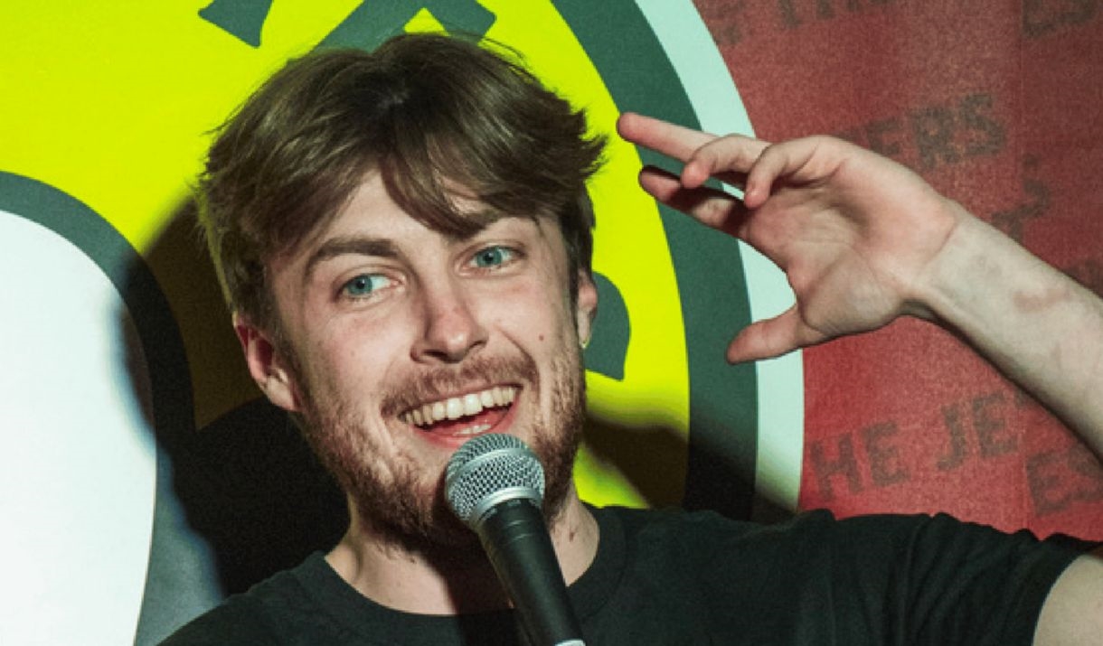 A young man doing stand up comedy. Sparkfest logo in top corner