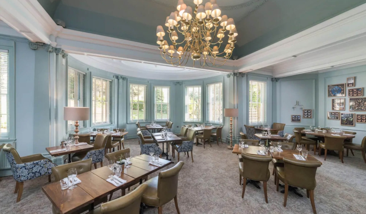 A horizontal view of the restaurant with soft chairs, wooden tables and a chandelier hanging above.