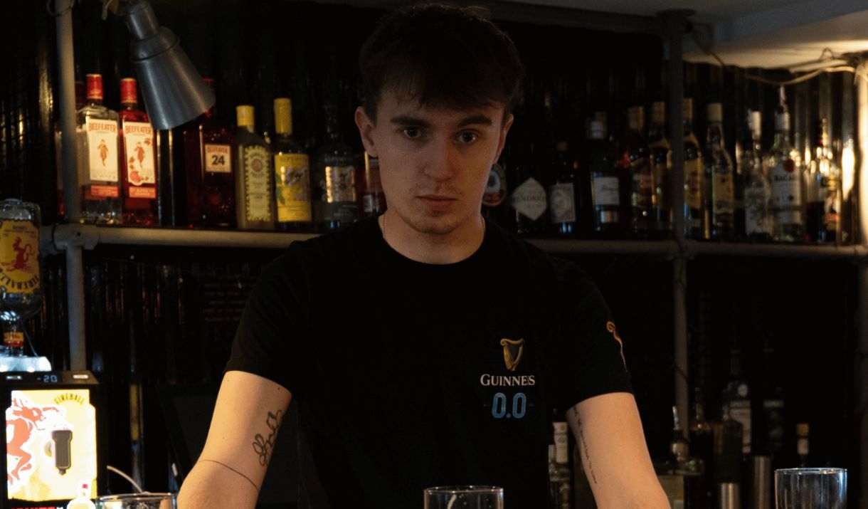 A young man standing behind a bar, staring, with a load of empty pint glasses in front. Sparkfest logo in top corner