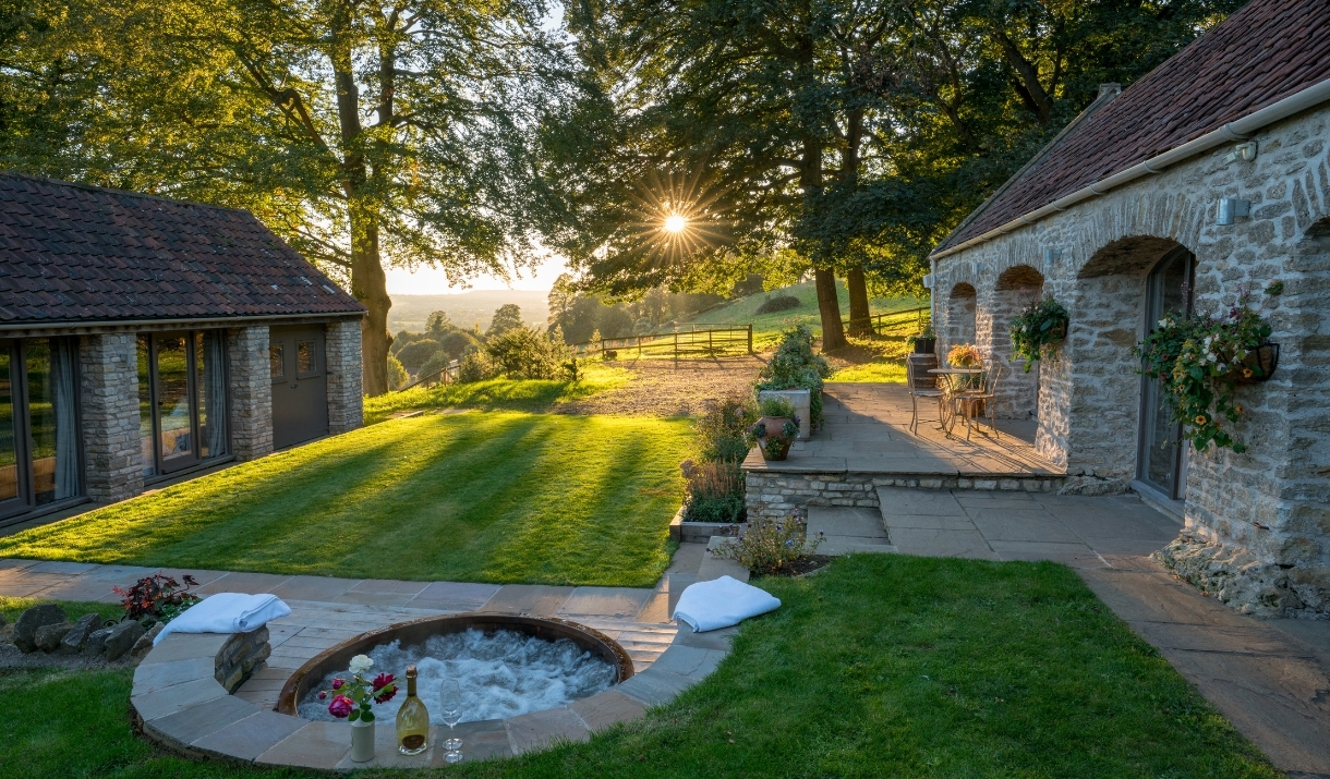 Scenic shot of a hot tub with champagne, the sunny countryside in the background, and the two brick lodgings.