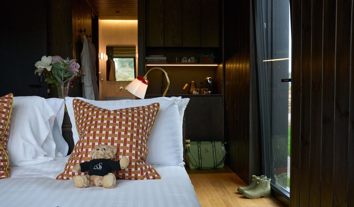 Close up of bed with nice brown cushions on white linen with a teddy bear on it and flowers and Barbour wellies in the background.