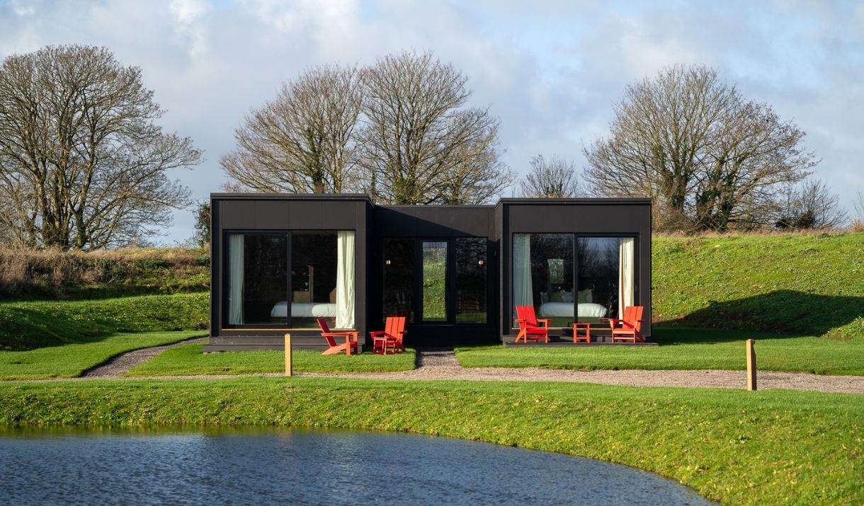 Modern-style black and white accommodation building with red chairs in front, overlooking a lake.