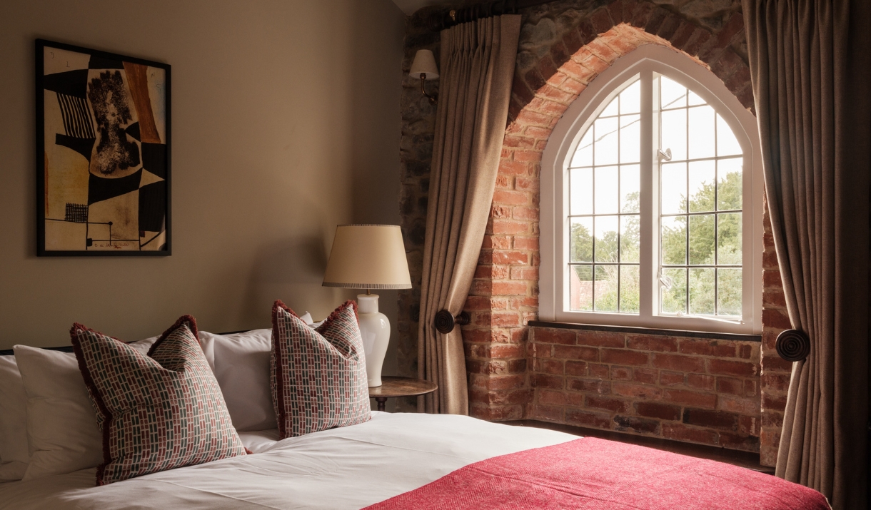 A double bed alongside an exposed brick wall with a large arched window and white curtains.