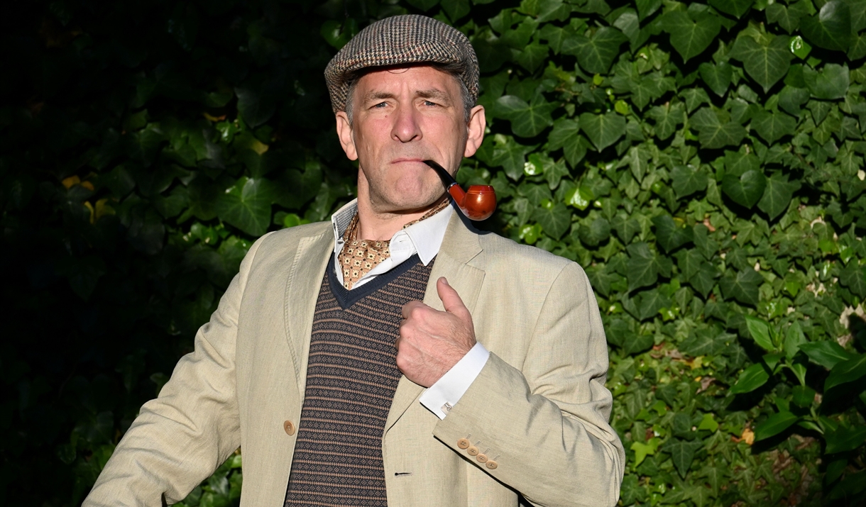 Matt Nation stands heroically, pipe in hand, as the show's hero Hugh 'Bullshot' Crummond. He is dressed in cream linen suit, tweed flat cap and fairis