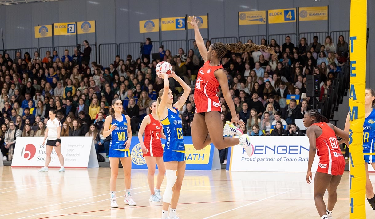 A defender, wearing red, leaps into the air as a Team Bath Netball shooter, wearing blue, lines up a shot. There is a stand of spectators watching beh