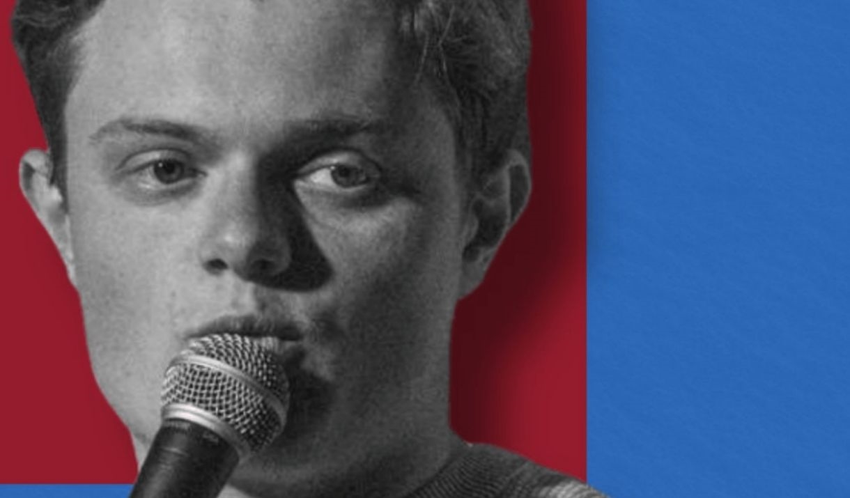 A young man (with a somewhat square head) doing stand up comedy. Sparkfest logo in corner