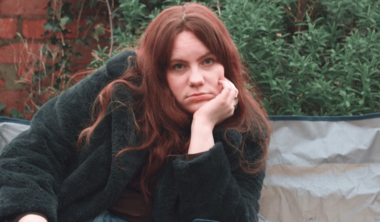 A girl slouching on a picnic chair, the chair next to her is empty except for a cupcake with a singular candle. Sparkfest logo in top corner.