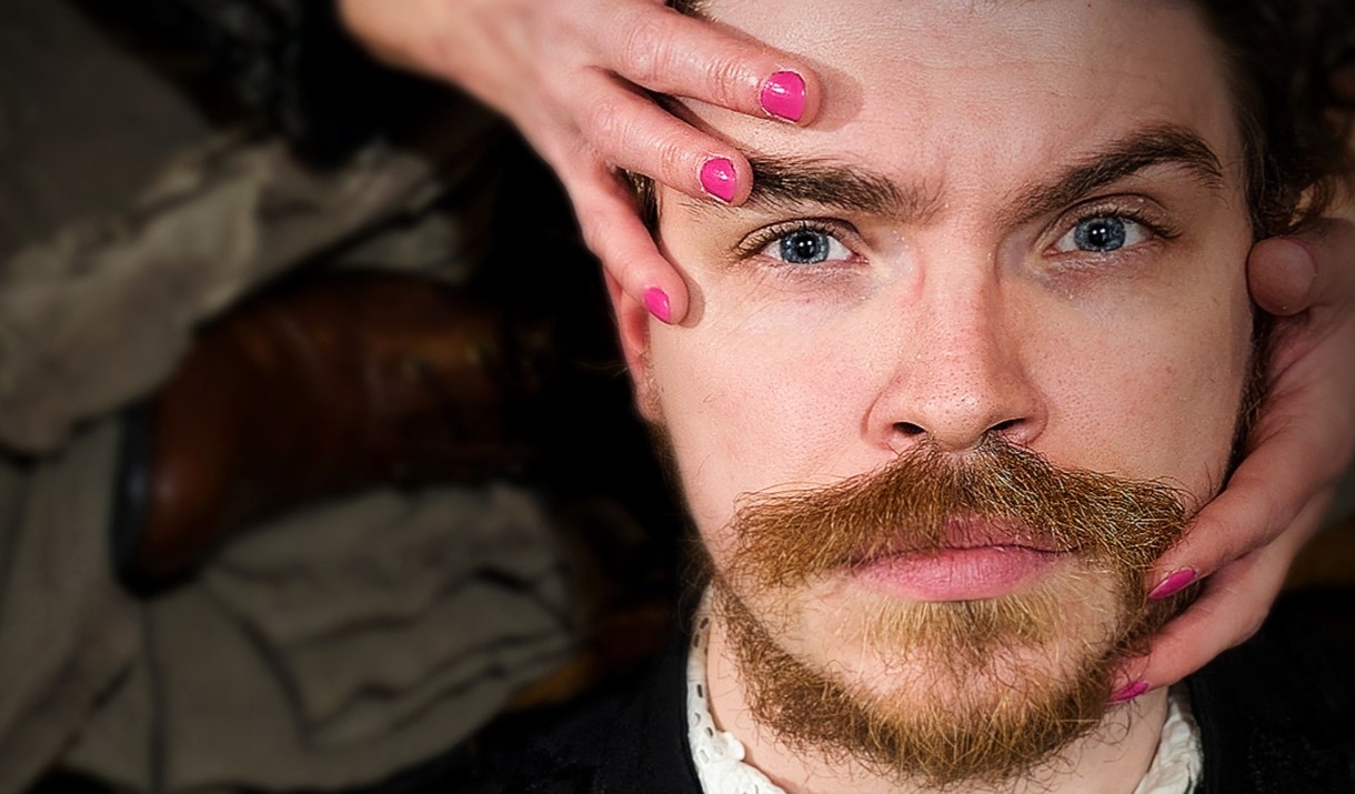 A white man with facial hair stares directly into the camera, while two hands with fingernails painted pink clasp his face.