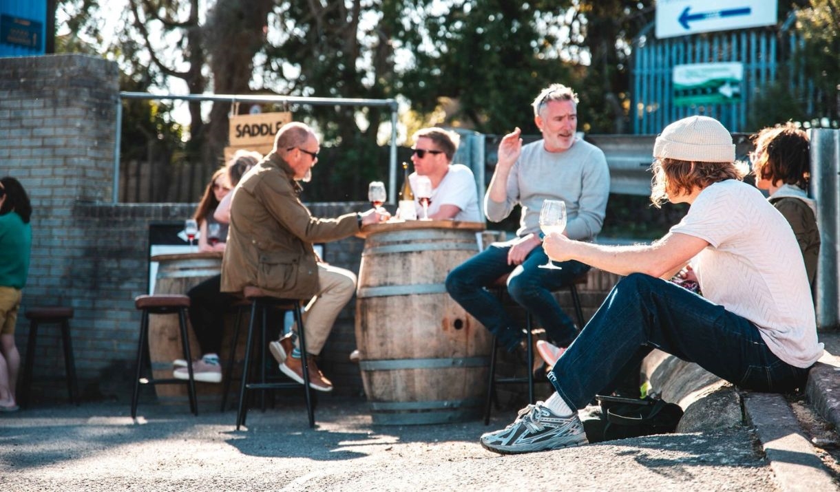 Guests at Saddle Goose's Cellar Door sat outside drinking from wine glasses on a sunny day. Some patrons are sat on bar stools while others are seated