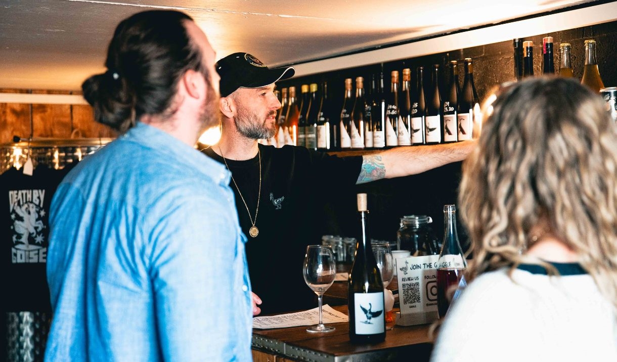 Adam at Saddle Goose, wearing a black t-shirt and cap, points at a row of wine bottles on a shelf while serving two customers.