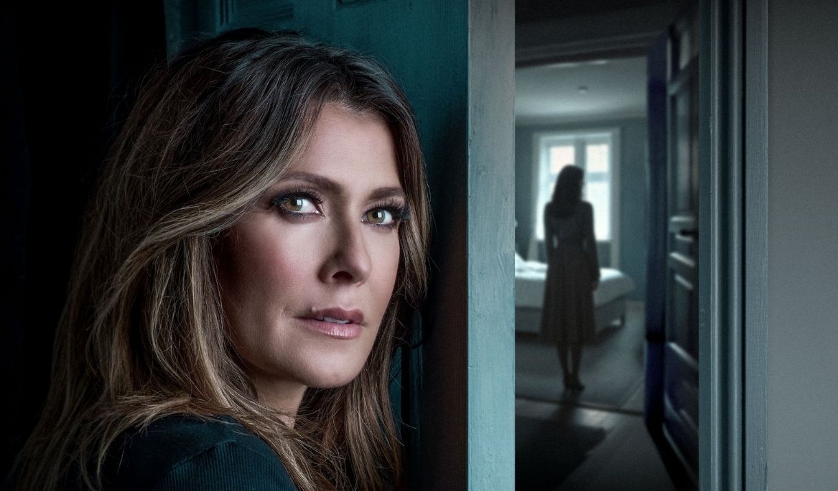 A woman stands behind a door, with a mysterious woman in a white dress in the background, not facing the camera.