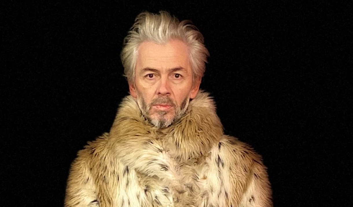 A man stands in front of a black background, he wears a large animalistic coat.