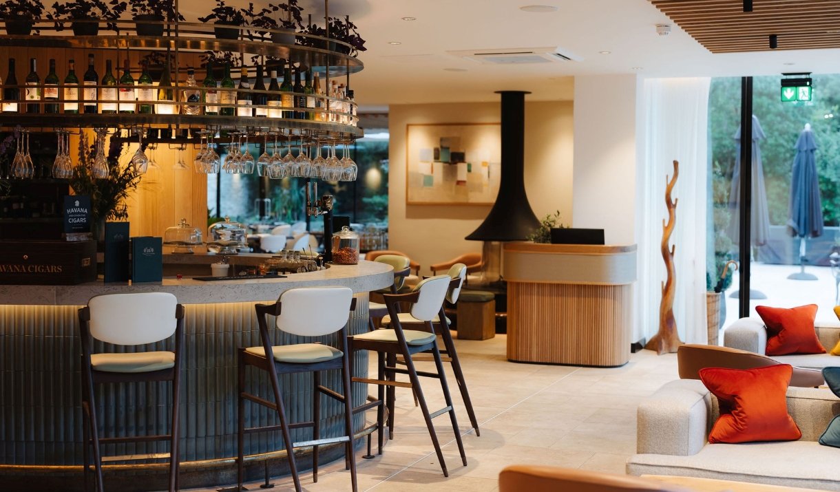 A smart indoor bar and seating area with white and black bar stools around a curved black panelled bar with white sofas to the right and a floor to ce