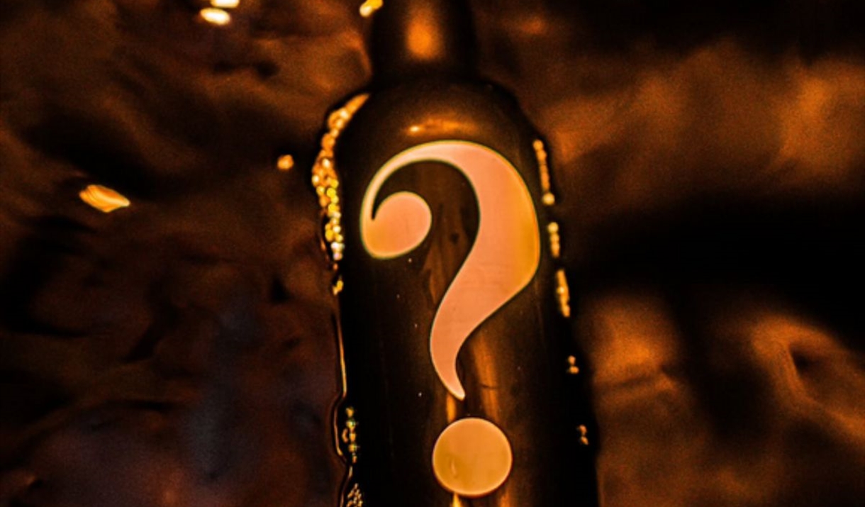 A black whisky bottle with a question mark on it. The background is also dark with smoke and embers of fire floating around the bottle.