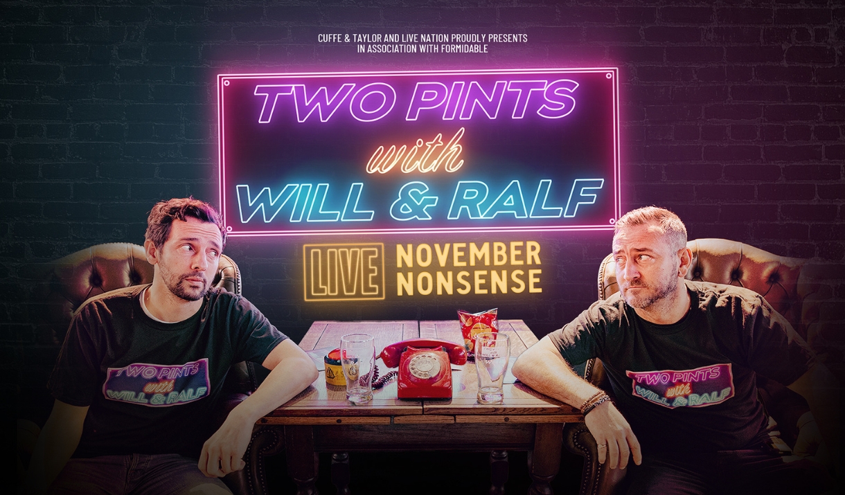 two men sitting either side of a table looking at one another, with a telephone and two pint glasses on the table