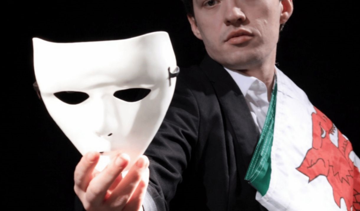 A man wearing a suit with a folded Welsh flag on his shoulder and holding out a plain white theatre mask. Sparkfest logo in top corner