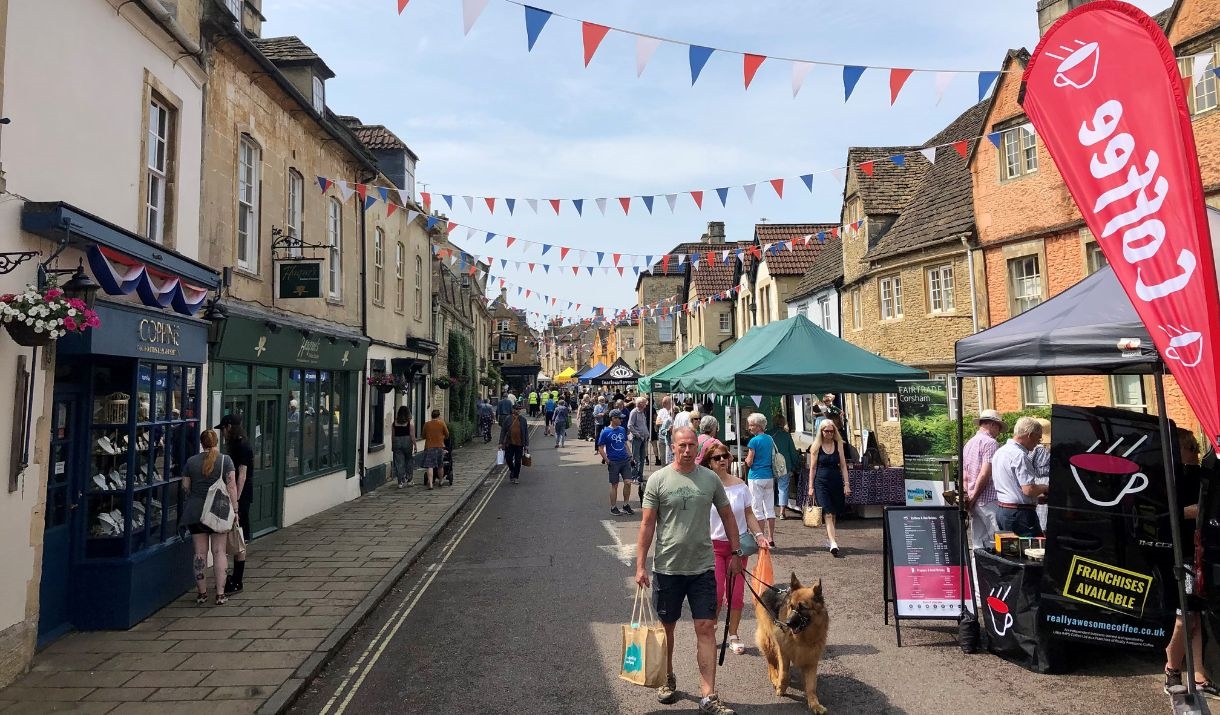 A view of Corsham's high street, with old buildings and food and drink stalls along the roads. There are lots of people browsing the stalls and walkin