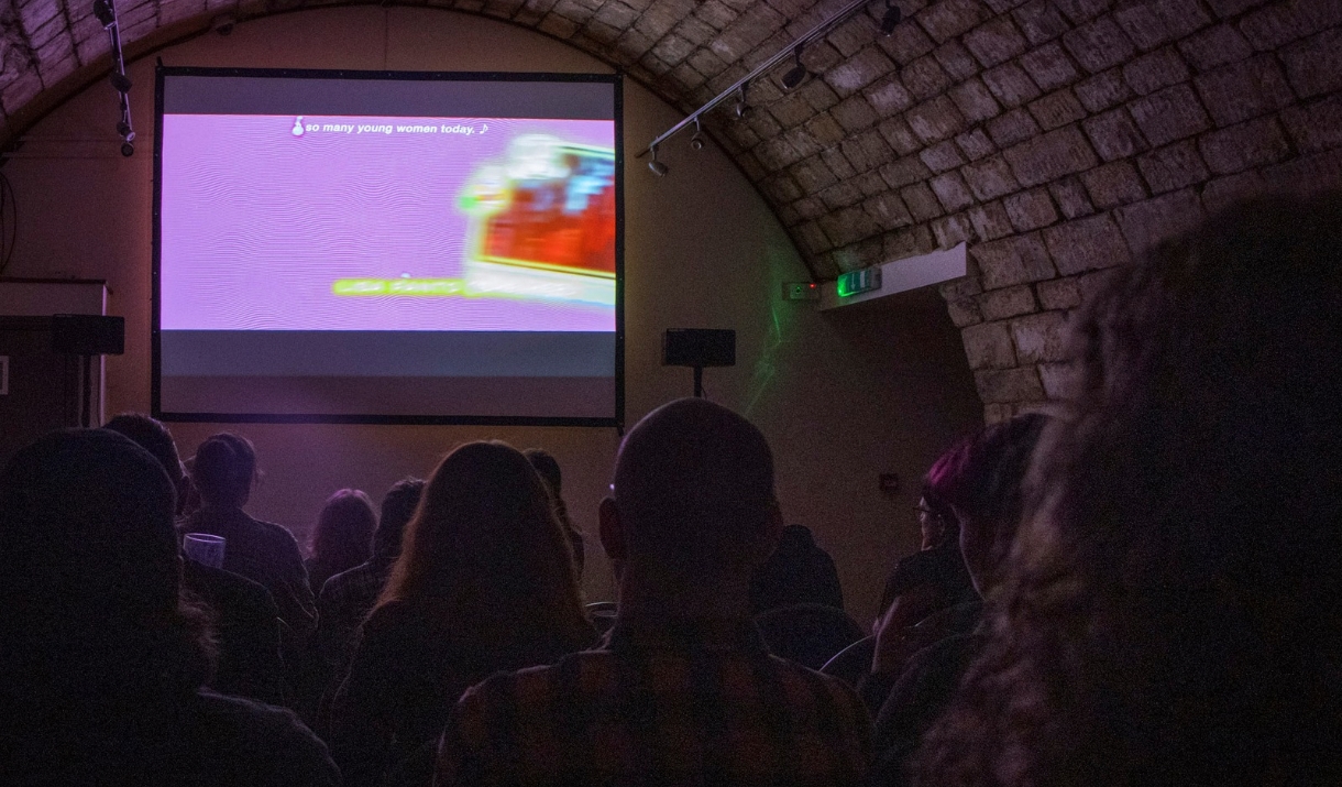 A purple-lit image of a groups of people facing a large screen, showing T-Blockers, a film directed by Alice Maio Mackay.