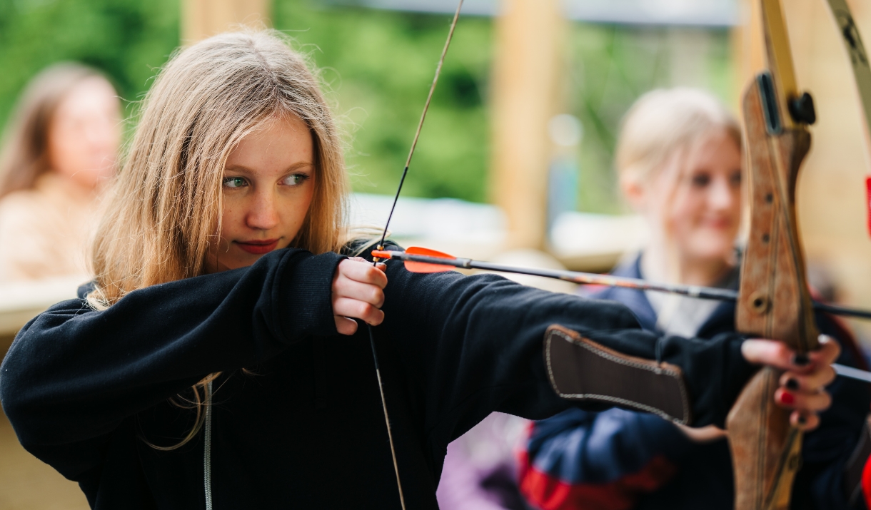 Teenage girl with blonde hair, wearing a black hoodie, pulling back an arrow on an archery bow. Looking straight down the arrow with determination.