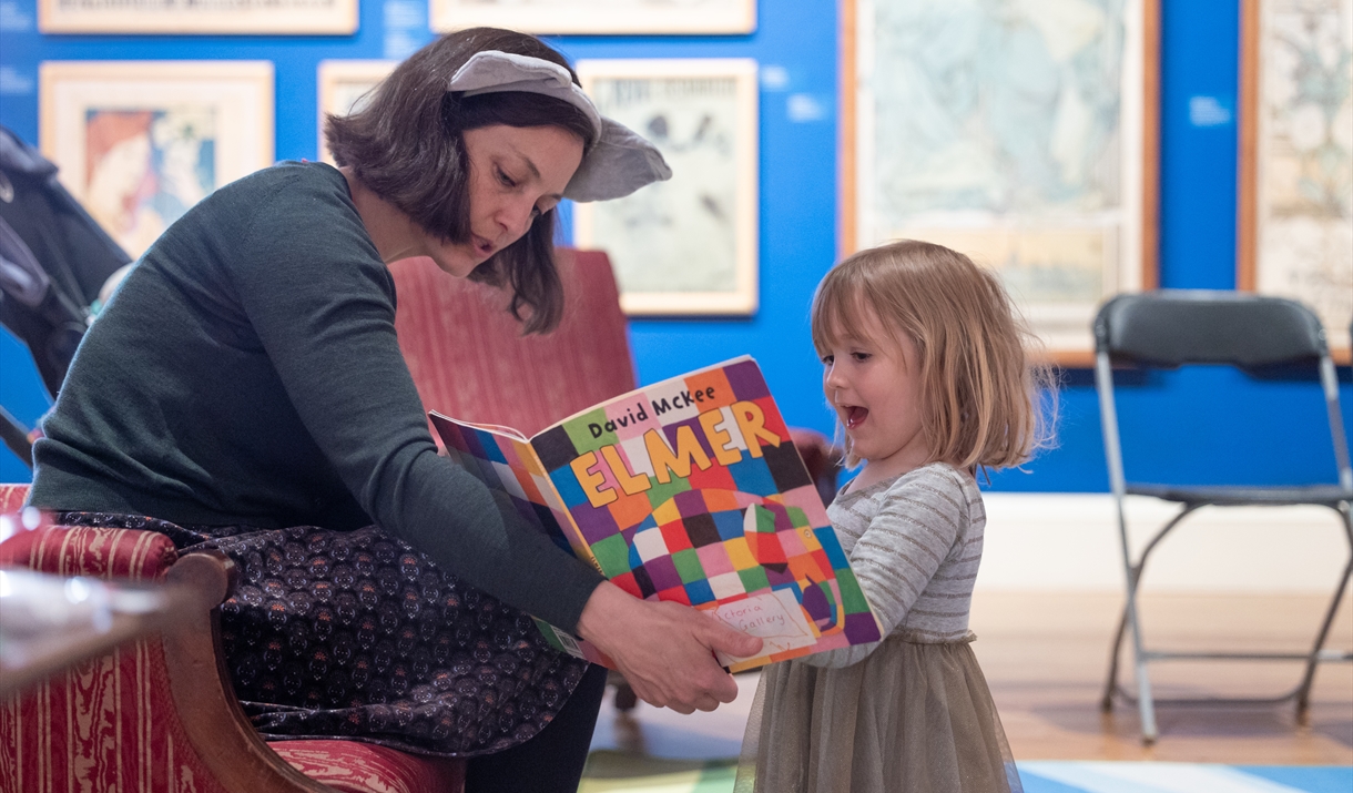 A lady reading wearing a headband with furry ears, reading a book to a young girl.