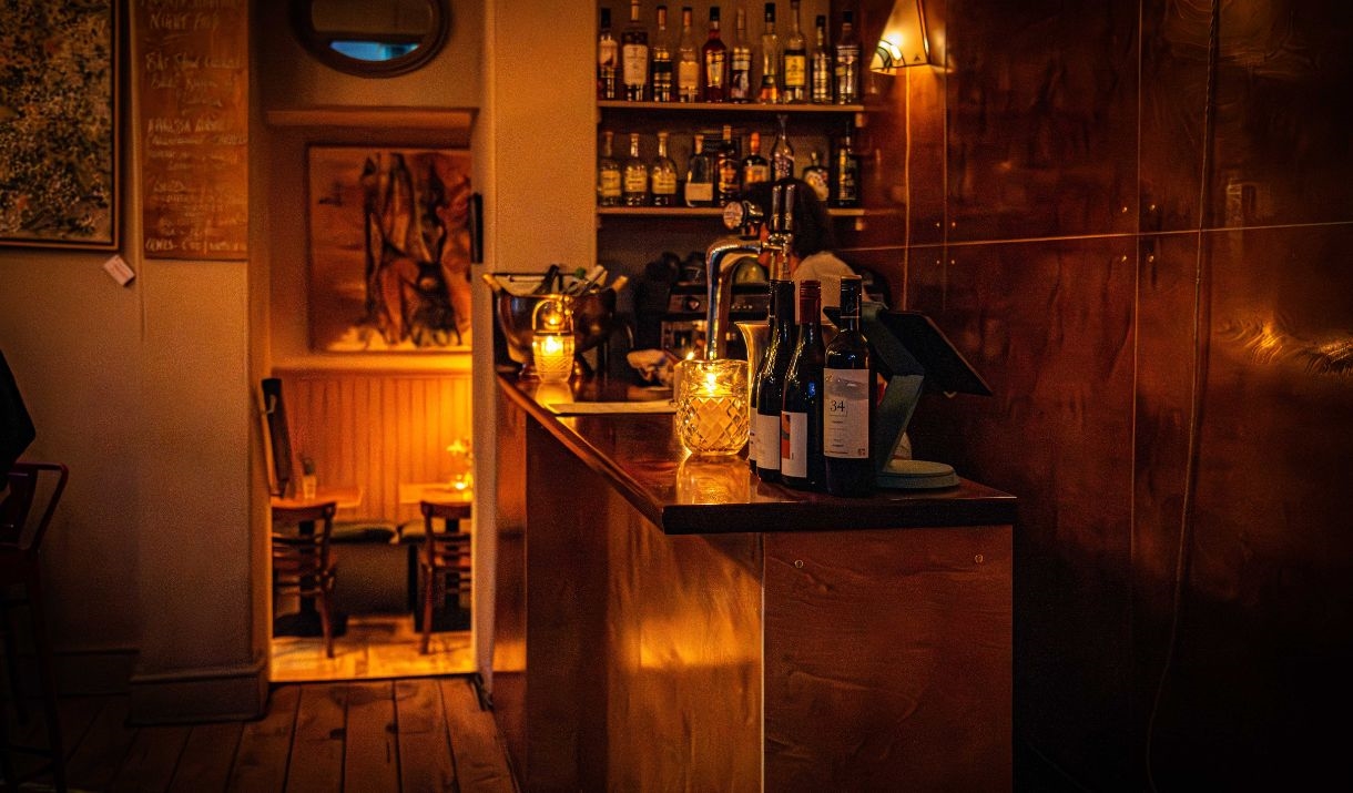 A side view of a bar containing several bottles of wine with warm lighting. Two shelves containing alcohol bottles are visible behind.