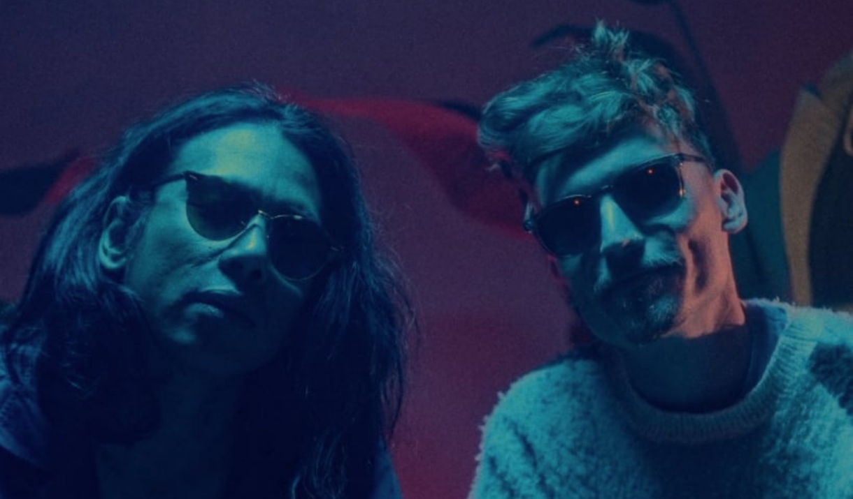 two men sit with sunglasses on, bathed with a blue light