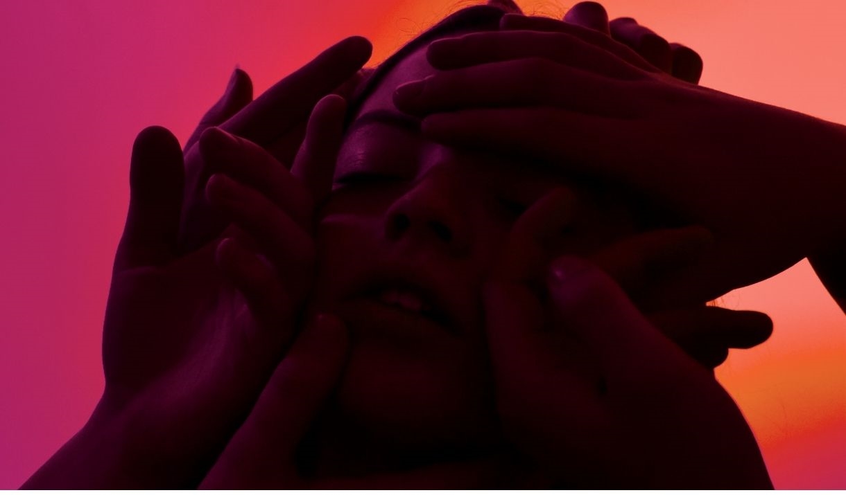 Close-up of a person's face surrounded by several hands, with dramatic red-pink lighting creating a moody, intense feel.