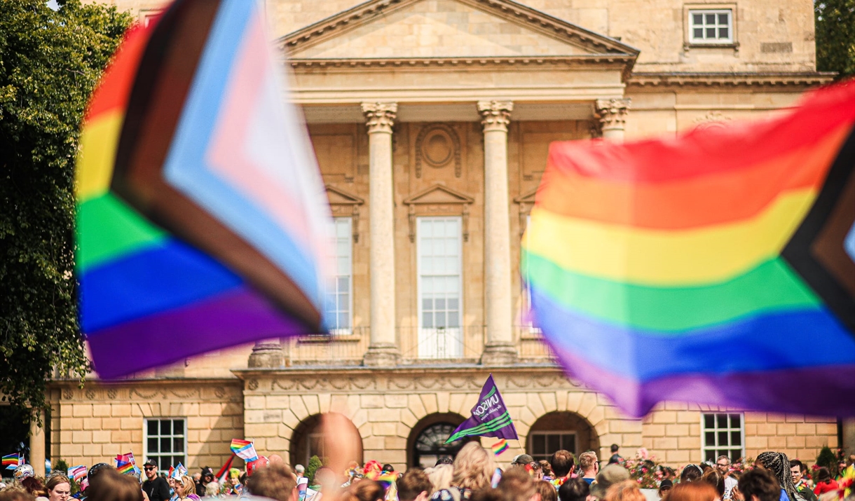 A photograph of the front of the Holburne Museum with a crowd of people in front. Two large progress rainbow flags fly in the foreground.
