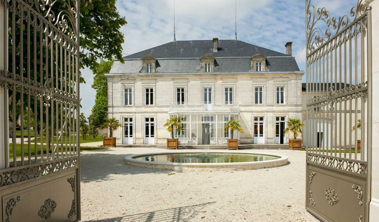 An image of the gated front to a 18th century french cognac chateau. The building is a large stone 3 floor chateau with a circular pool in front and a