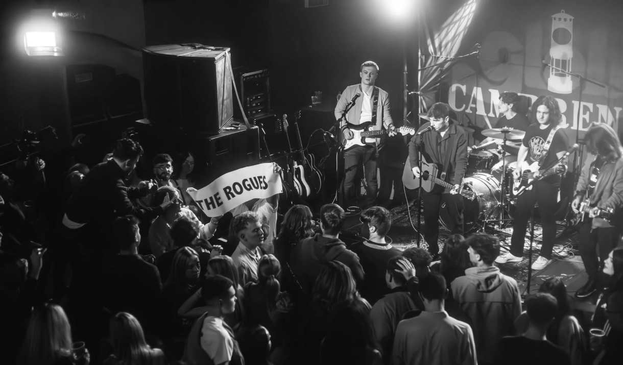 A black and white photo of a band playing live. A person in the crowed is holding up a banner with the words "The Rogues" written on it.