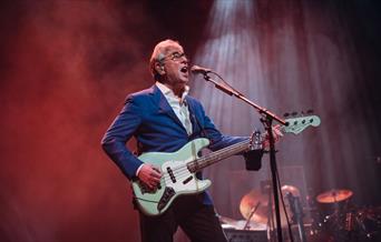 A photo of Graham Gouldman. He is playing an electric guitar while singing