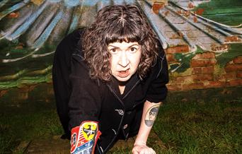 Amy Mason crawls towards the camera, she is on grass with a cowboy boot on her right arm