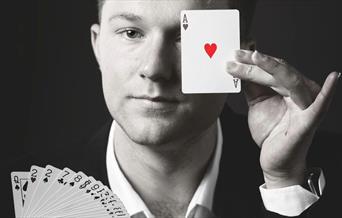 A black and white photo of the Magician Angus Baskerville, He is holding a deck of cards and covering one eye with the ace of hearts.