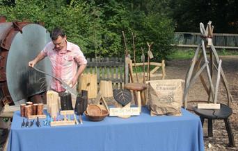 man standing in woodland by table displaying wooden craft objects