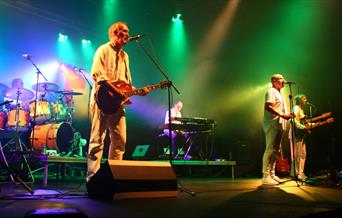 A photo of G2 Definitive Genesis performing on a stage. They are all wearing white clothing. Green, red, white and purple lights illuminate the stage.