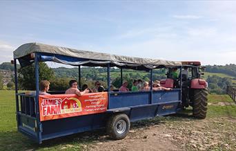 people sitting in a tractor trailer, in a green field, on a sunny day with an orange Jamie's Farm banner on the side of the trailer