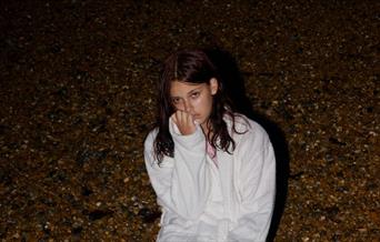 It's evening, a young woman in a white dressing gown sits down on a beach.